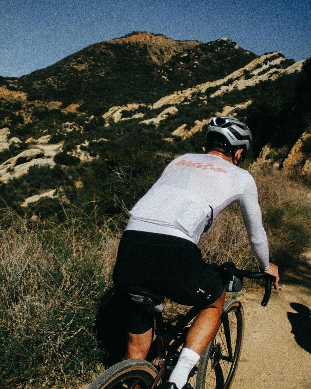 Cyclist riding a bicycle on a trail near a mountain with sparse vegetation under a clear sky, creating a sense of outdoor adventure.