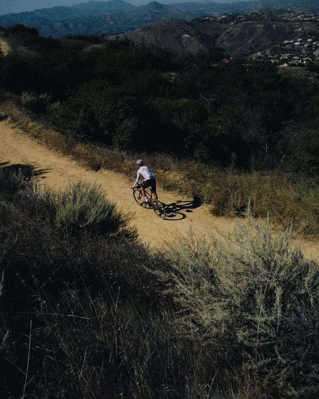 Cyclist riding a mountain bike on a dirt trail in the hills, surrounded by dry brush and trees under bright daylight.