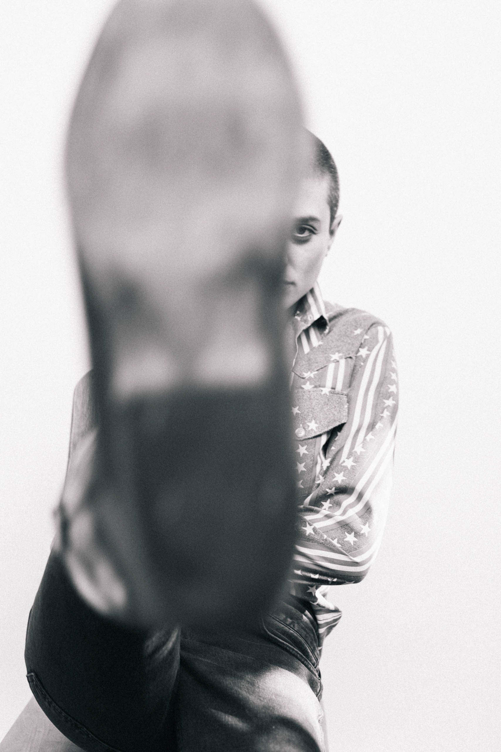 A monochromatic, high-contrast studio portrait of a woman with short hair in an American flag shirt and a large boot in the foreground.