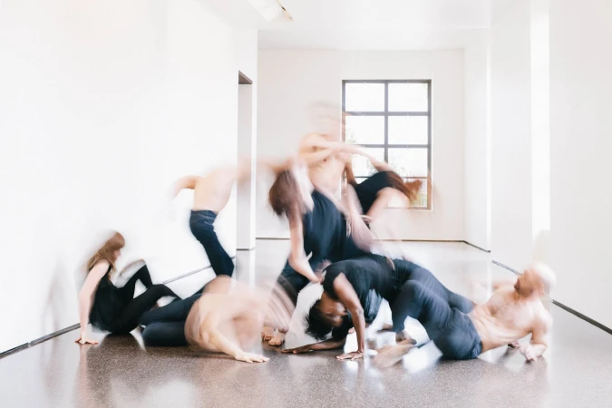 Group of dancers in motion, performing in a modern, bright white studio with a large window, captured with intentional motion blur effect.
