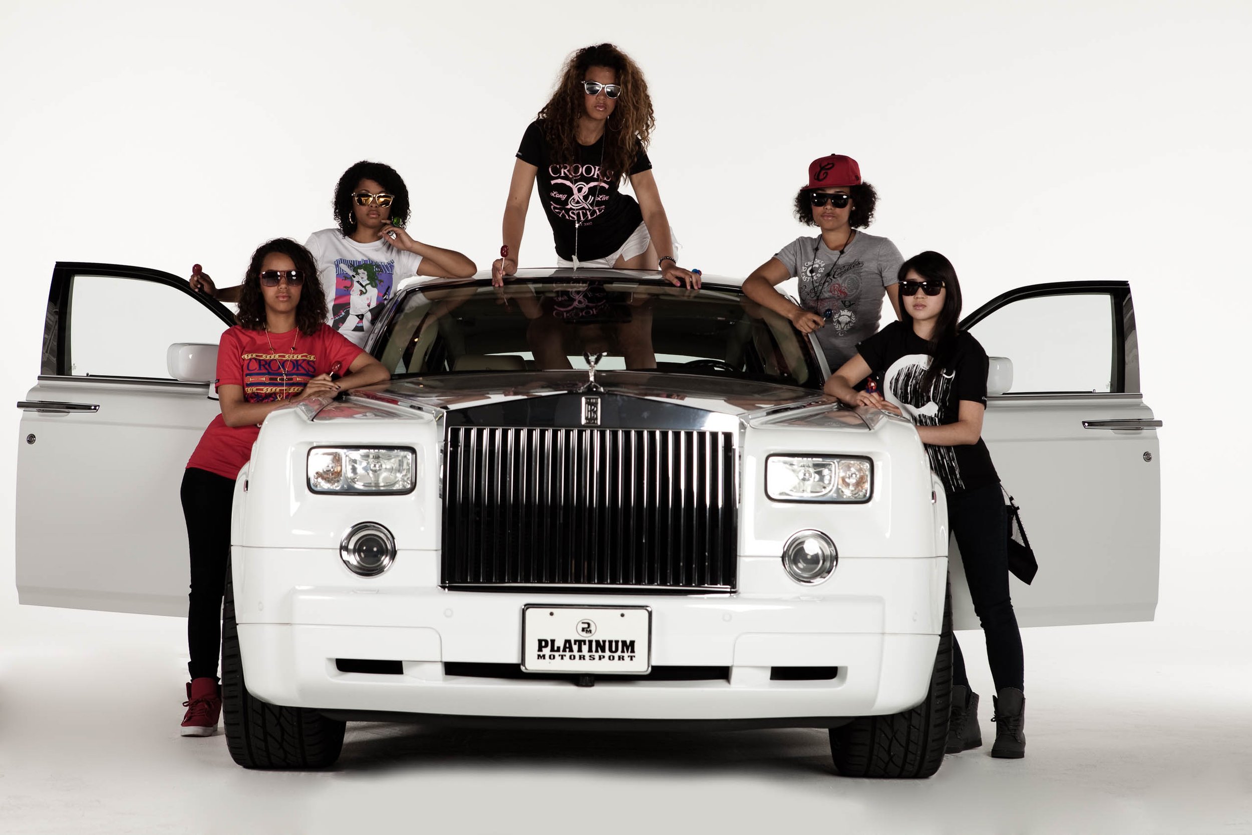 Five stylish women pose with a white Rolls Royce in a studio setting. They wear fashionable casual clothing and sunglasses, doors open.