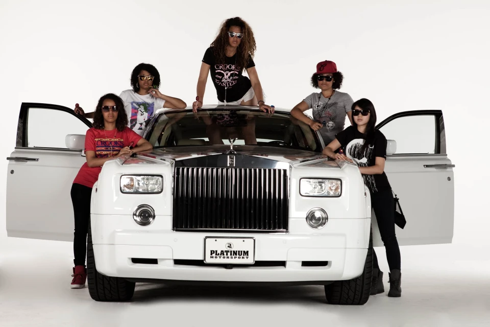 Five stylish women pose with a white Rolls Royce in a studio setting. They wear fashionable casual clothing and sunglasses, doors open.