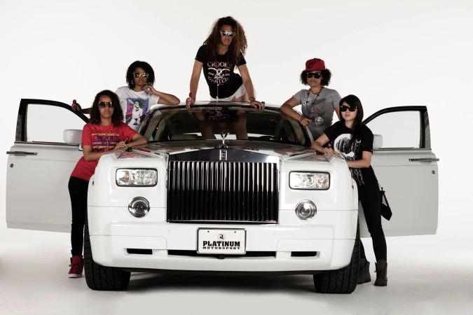 Five stylish women pose with a white Rolls Royce in a studio setting. They wear fashionable casual clothing and sunglasses, doors open.