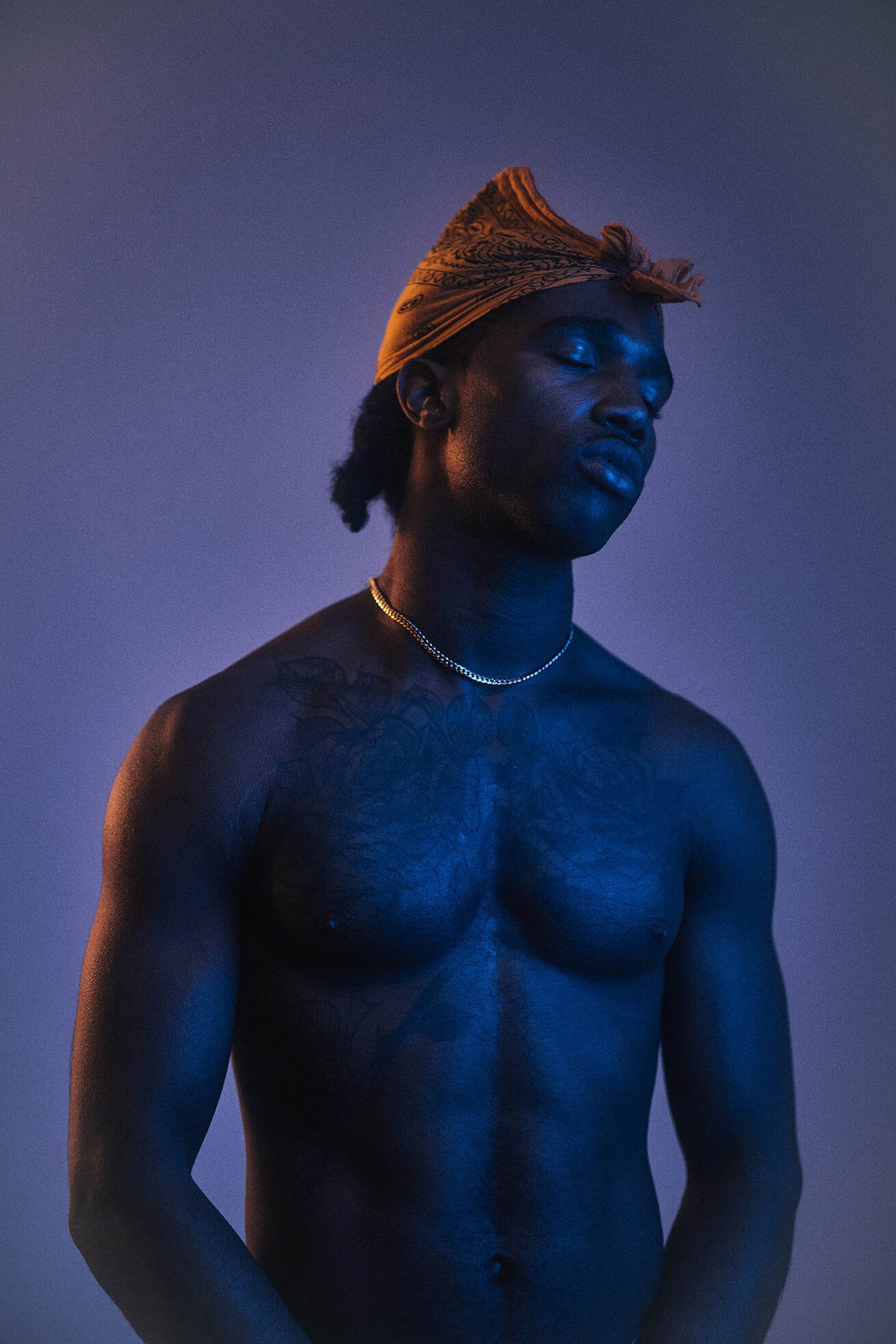 Striking portrait of a shirtless Black man wearing a gold chain and patterned headscarf against a cool-toned blue and purple backdrop.