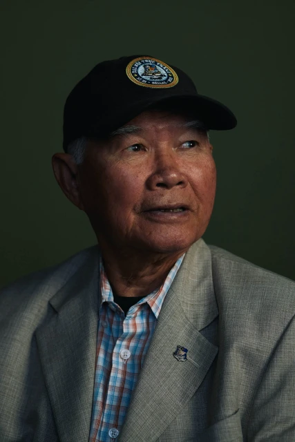 Portrait of an elderly Asian man wearing a cap, a patterned shirt, and a blazer against a dark green background.