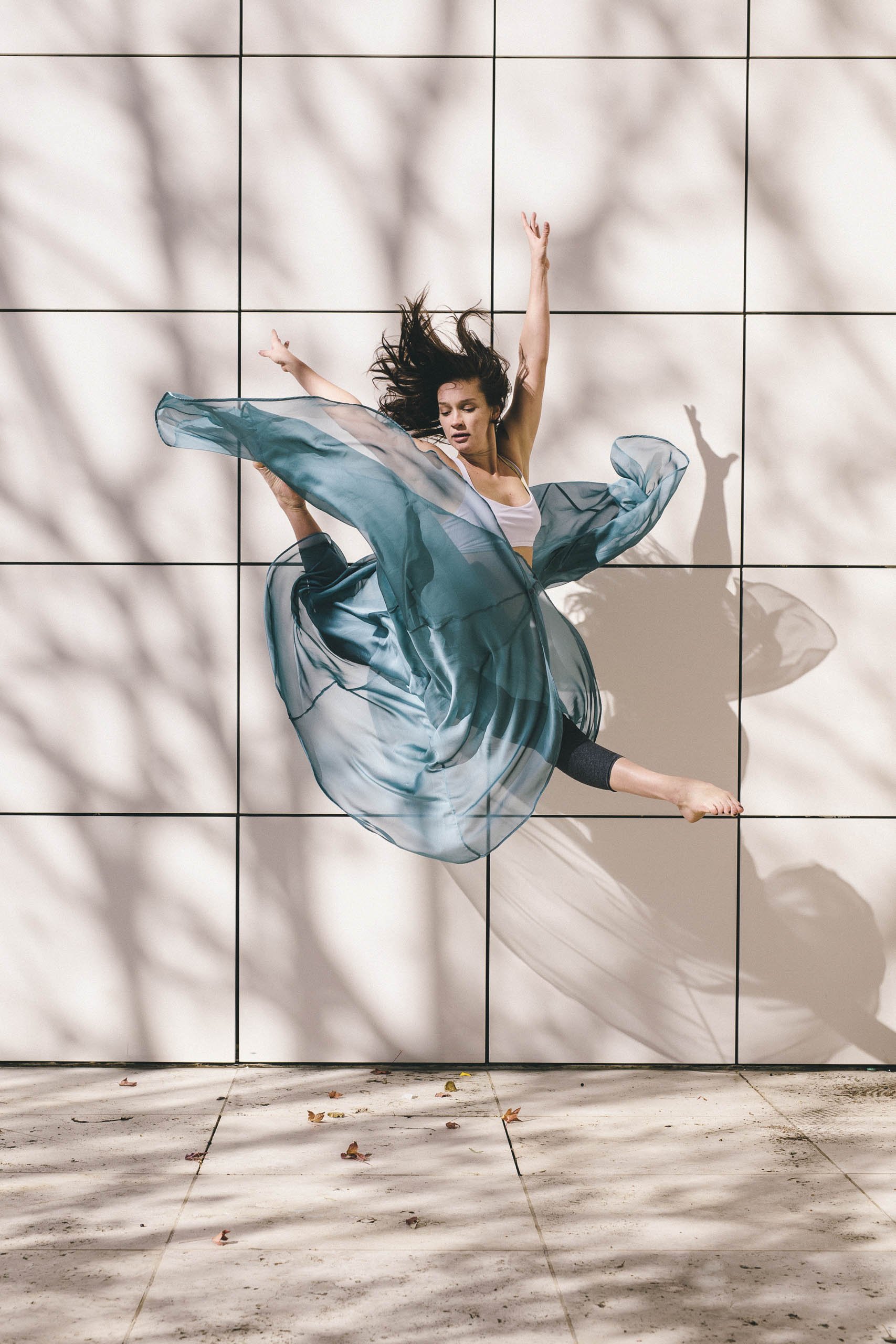 A woman in an athletic outfit with a flowing skirt leaps in the air in front of a tiled wall with shadow patterns on the concrete ground.