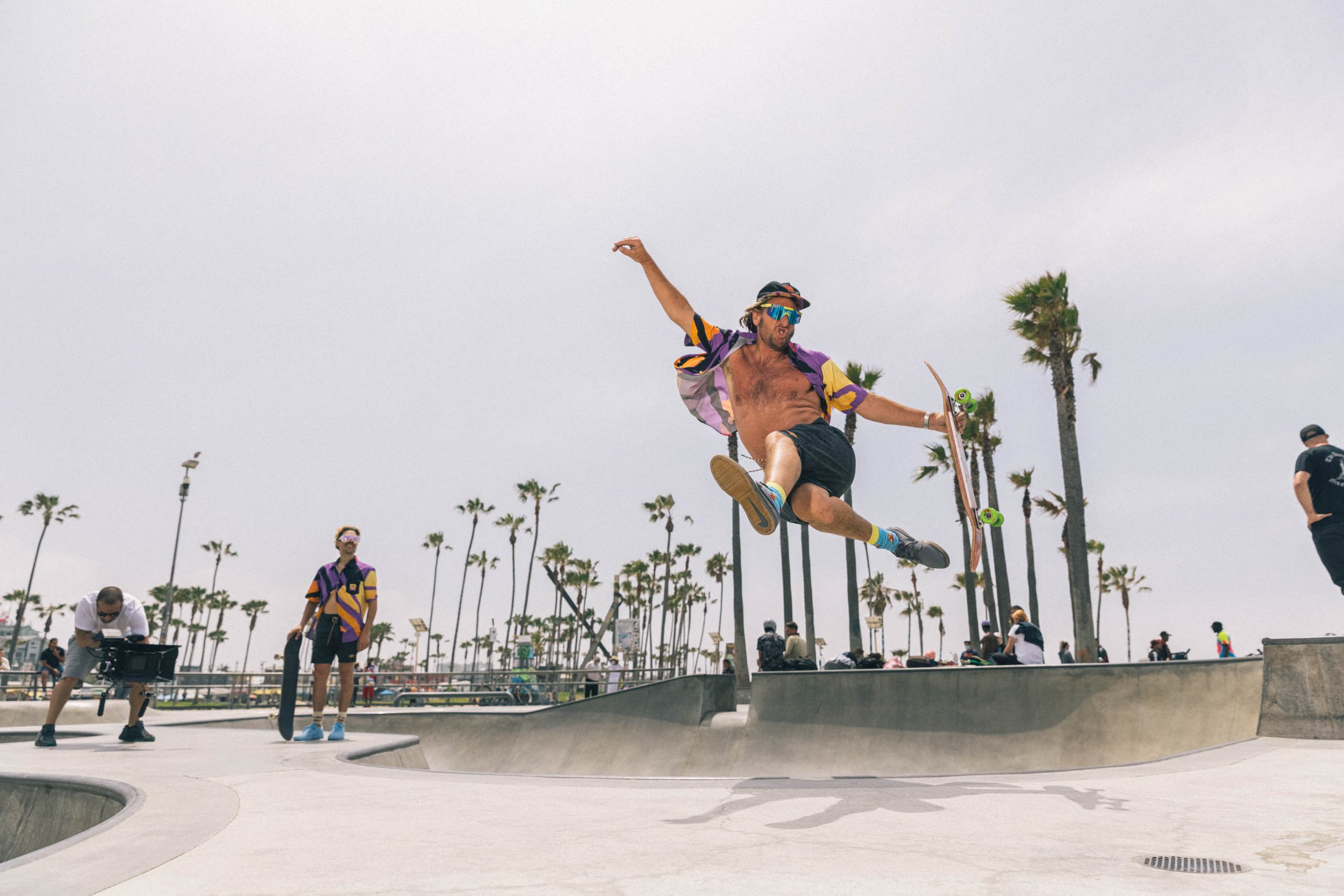 Man performs skateboard stunt at outdoor park, leaping high with board in hand, wearing sunglasses and a patterned shirt against palm trees.