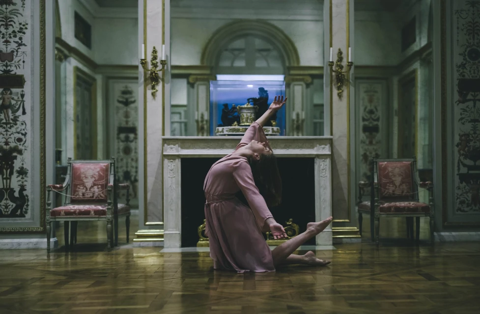 A woman in a light pink dress is kneeling on a reflective wood floor inside an ornately decorated room with marble walls.