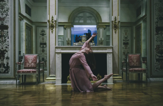 A woman in a light pink dress is kneeling on a reflective wood floor inside an ornately decorated room with marble walls.