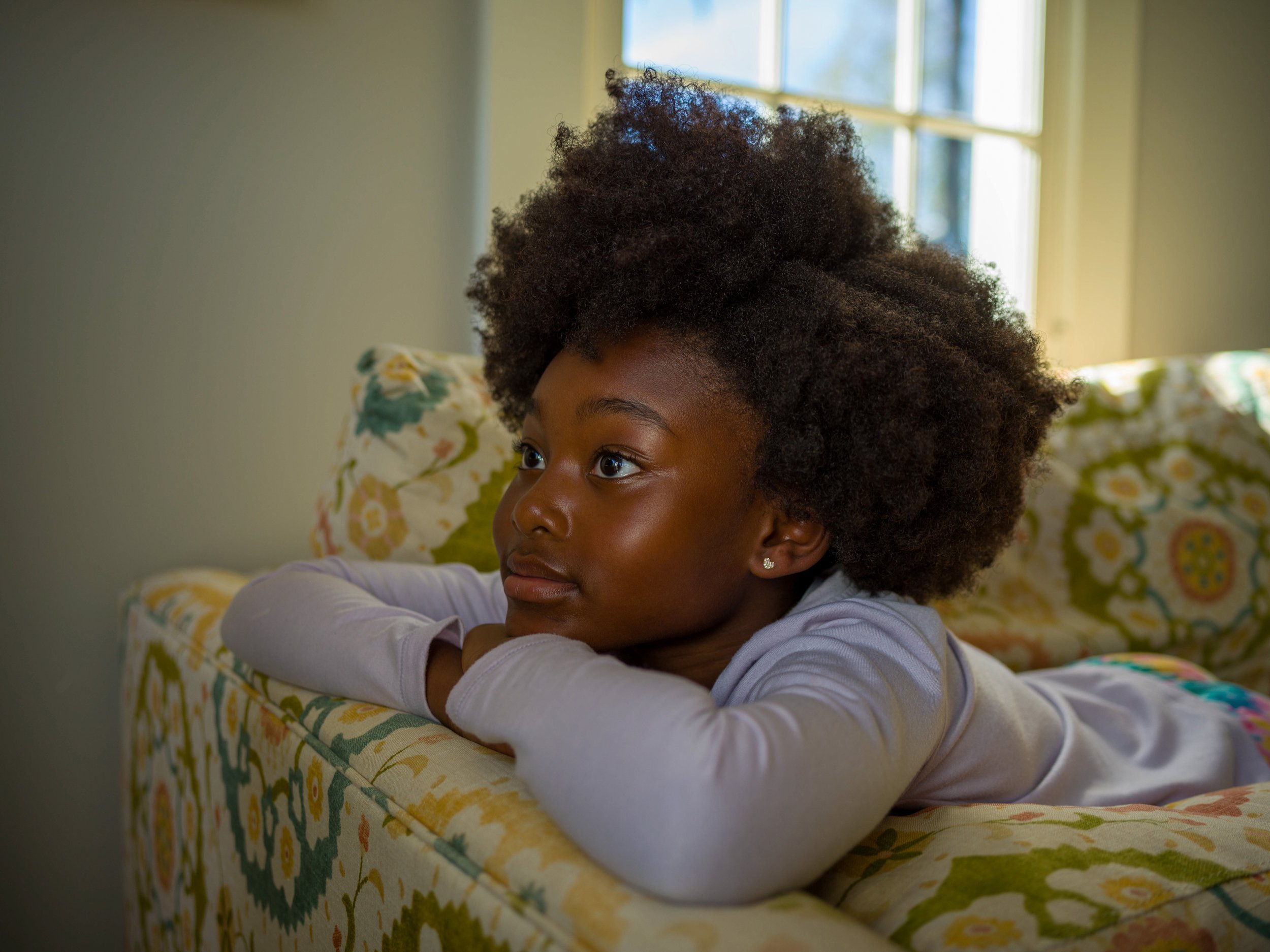 Young African-American girl with afro resting on a floral patterned sofa looking towards window with natural light coming through.