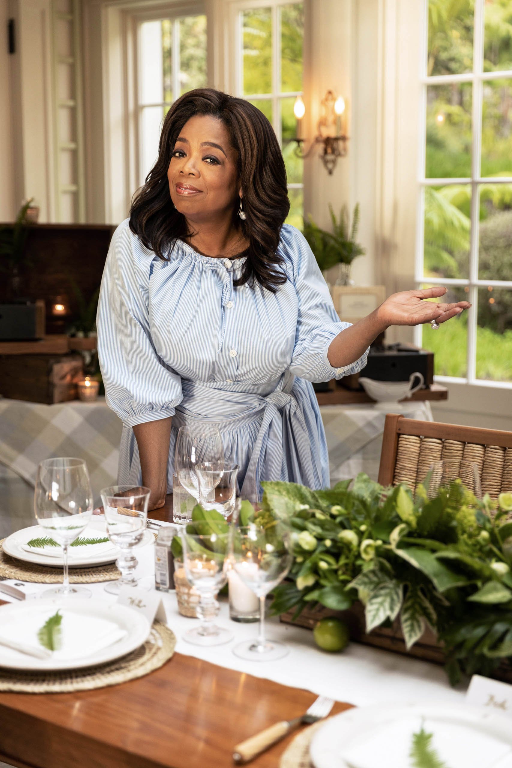 Oprah Winfrey smiling in a light blue dress standing at a beautifully set dining table with floral centerpieces