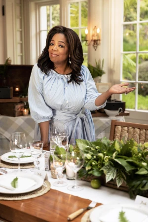 Oprah Winfrey smiling in a light blue dress standing at a beautifully set dining table with floral centerpieces