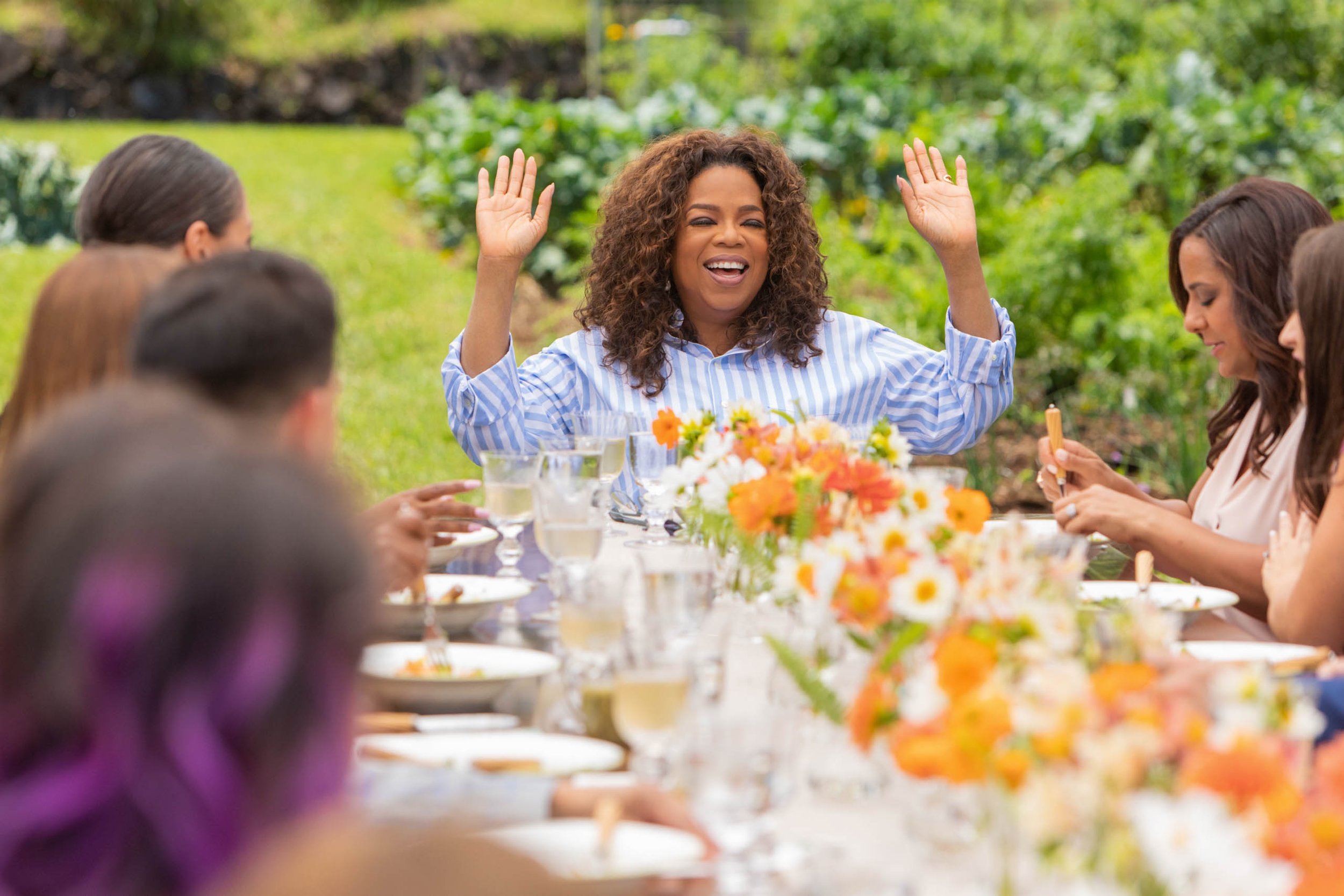 Oprah Winfrey raises her hands at an outdoor dinner table surrounded by guests, featuring flowers, wine glasses, and lush greenery.