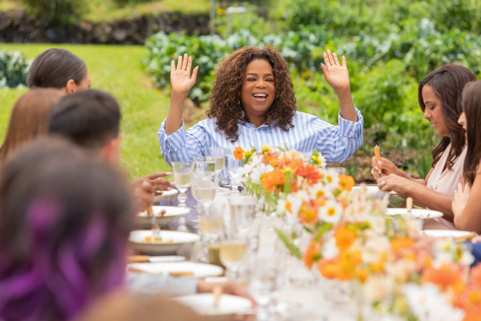 Oprah Winfrey raises her hands at an outdoor dinner table surrounded by guests, featuring flowers, wine glasses, and lush greenery.
