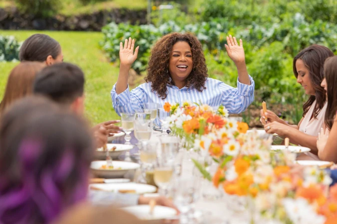 Oprah Winfrey raises her hands at an outdoor dinner table surrounded by guests, featuring flowers, wine glasses, and lush greenery.