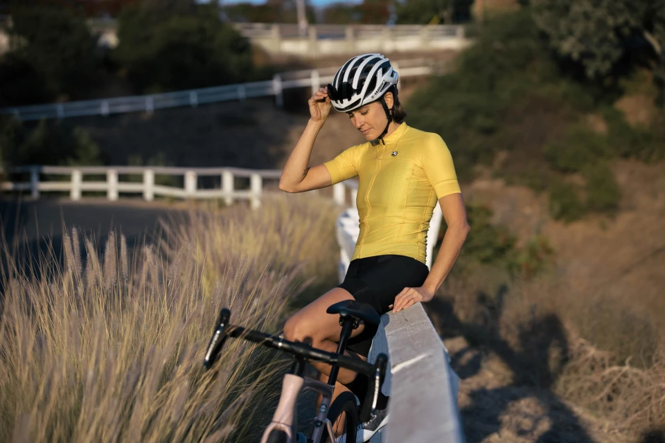 Female cyclist in yellow jersey and helmet rests on roadside barrier with bike, sunlit setting, outdoor, fitness, and cycling theme.