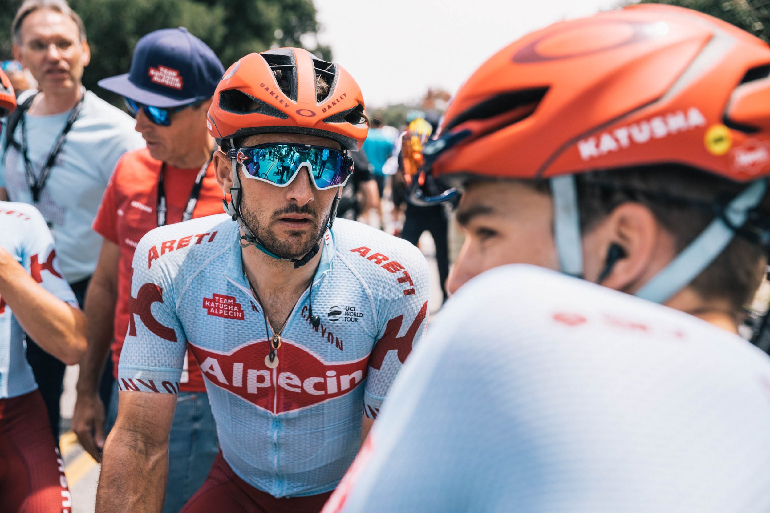 Two cyclists in team jerseys and helmets converse outdoors. The focus is on the cyclist wearing Oakley sunglasses and a light blue jersey.