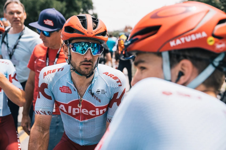 Two cyclists in team jerseys and helmets converse outdoors. The focus is on the cyclist wearing Oakley sunglasses and a light blue jersey.