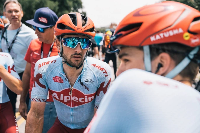 Two cyclists in team jerseys and helmets converse outdoors. The focus is on the cyclist wearing Oakley sunglasses and a light blue jersey.