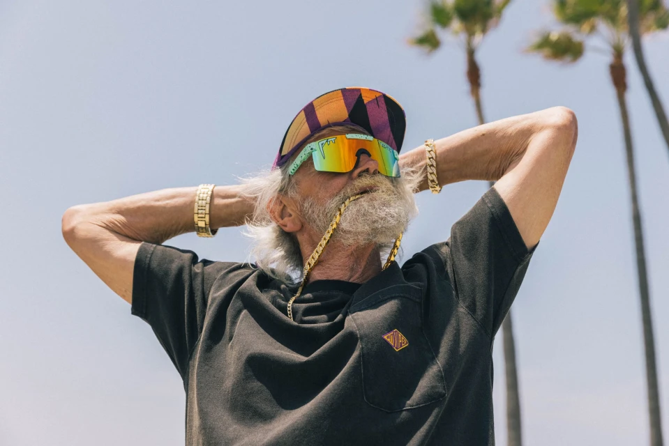 Elderly man with sunglasses and backwards cap poses with hands behind his head against a backdrop of tall palm trees on a sunny day.