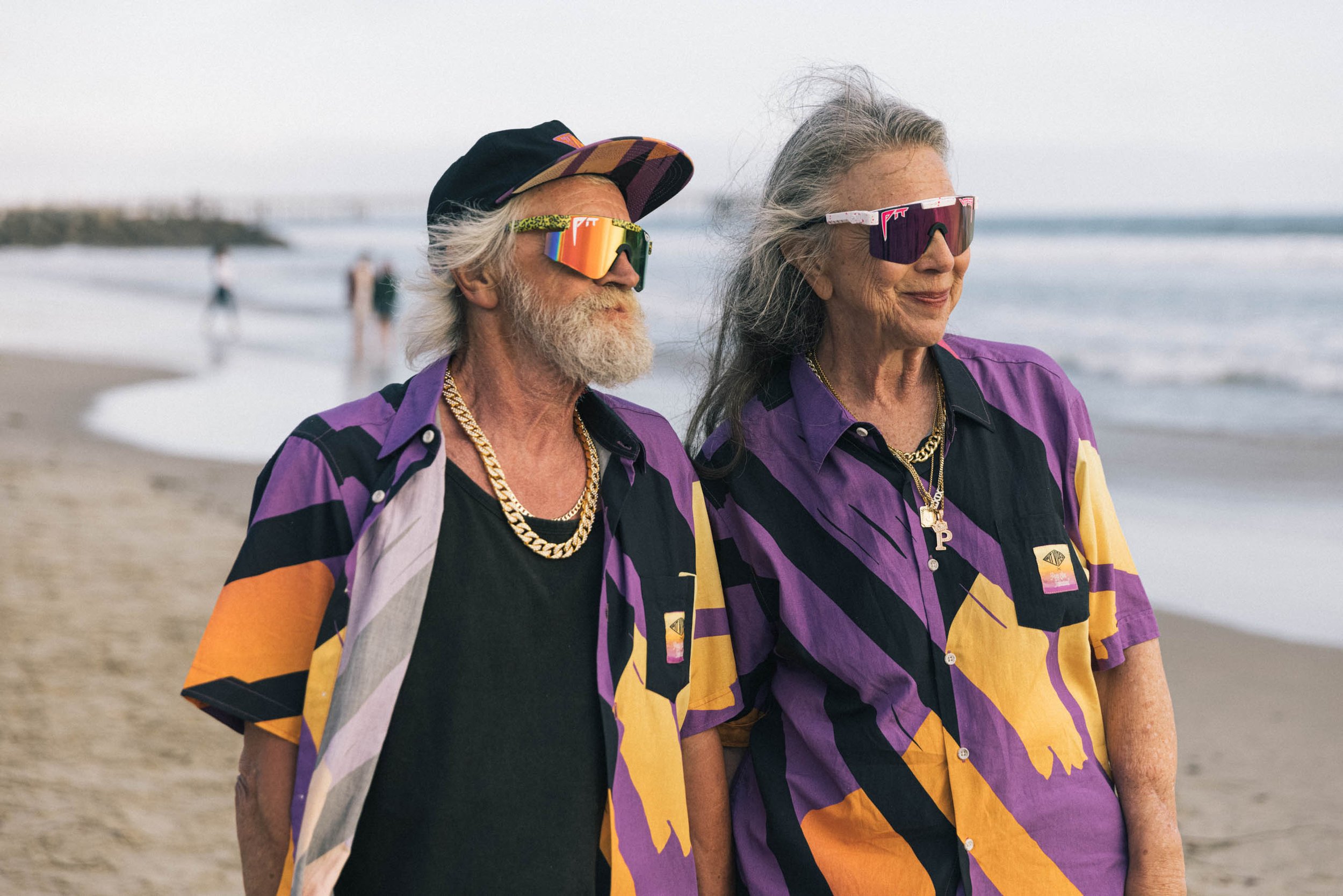 Elderly couple in retro sunglasses and colorful shirts standing on a sandy beach near the ocean on a sunny day with light surf.