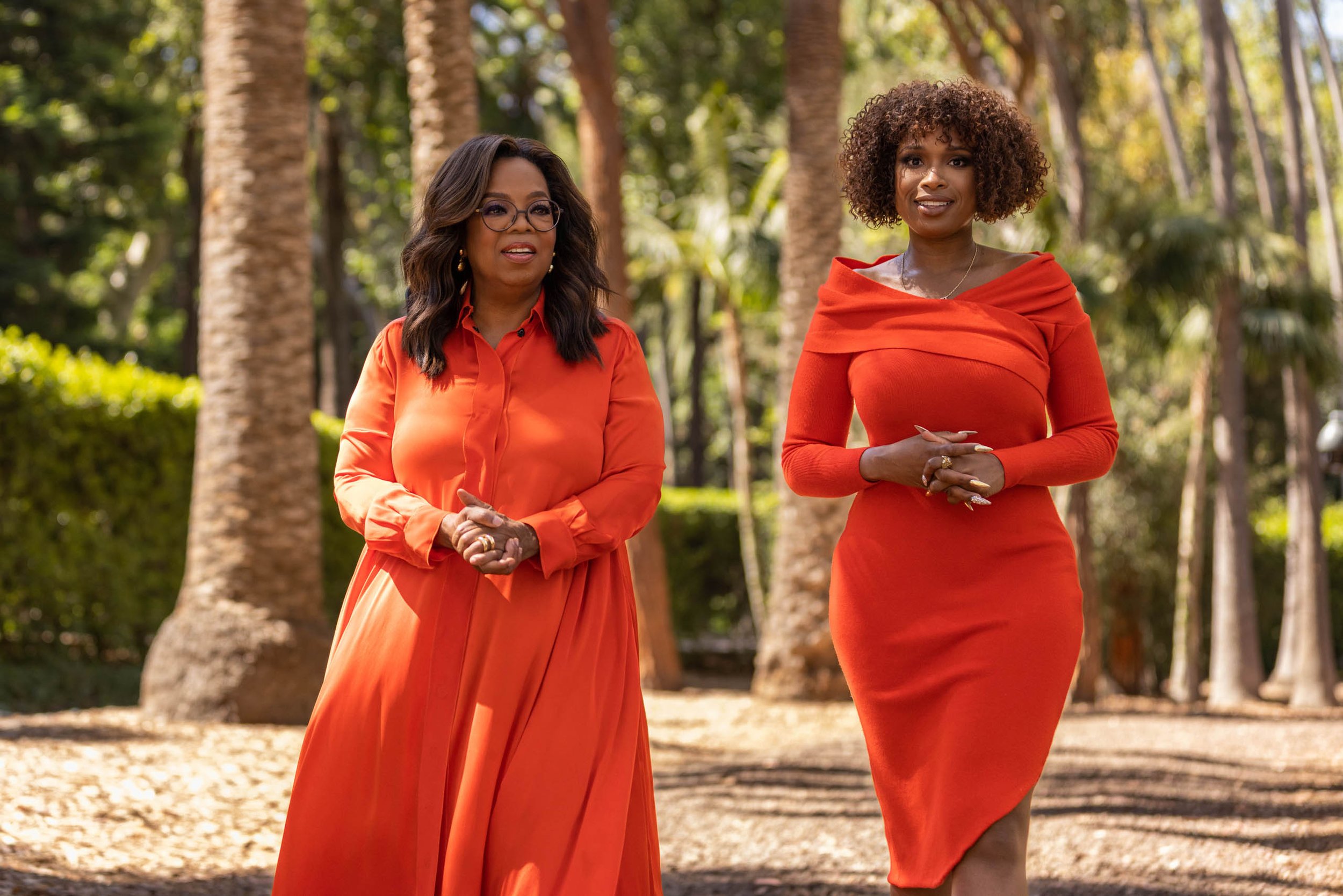 Oprah Winfrey and Jennifer Hudson pose outdoors in matching orange dresses amidst palm trees, creating a striking celebrity portrait.