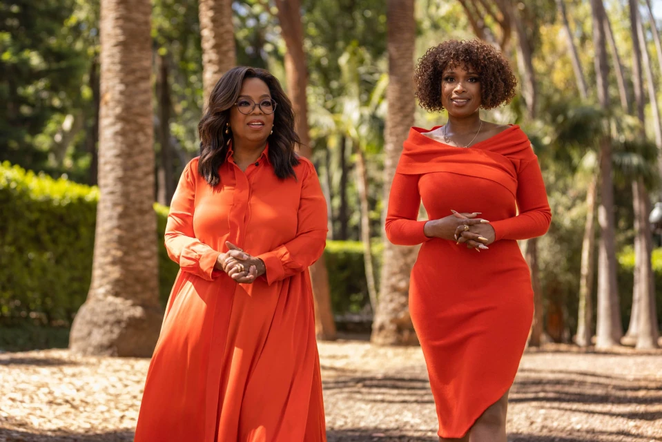 Oprah Winfrey and Jennifer Hudson pose outdoors in matching orange dresses amidst palm trees, creating a striking celebrity portrait.