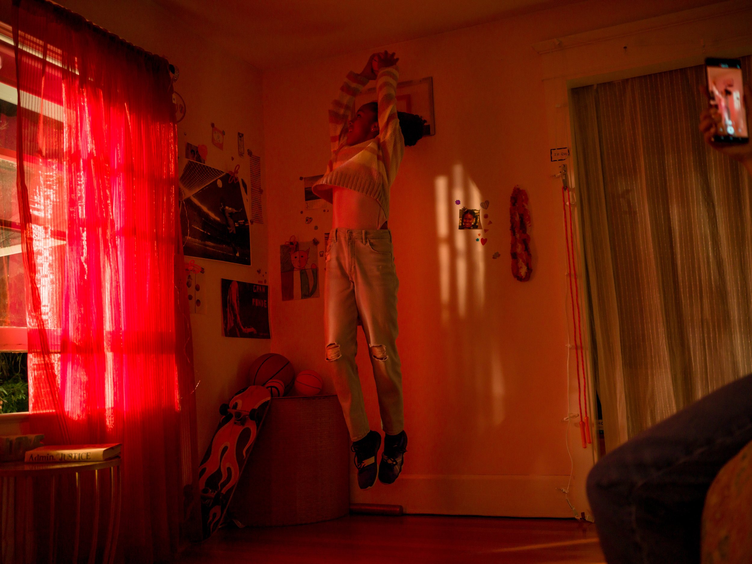 Teenage girl mid-jump in bedroom, phone recording visible. Red curtains, skateboard, and casual clothing fill the interior.