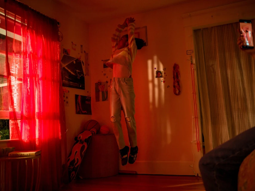 Teenage girl mid-jump in bedroom, phone recording visible. Red curtains, skateboard, and casual clothing fill the interior.
