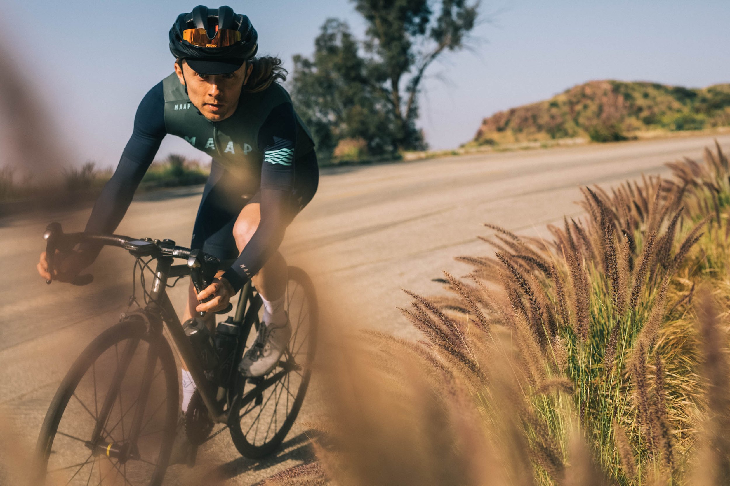 Cyclist in dark green jersey and black helmet riding road bicycle along a paved road past tall grassy weeds.