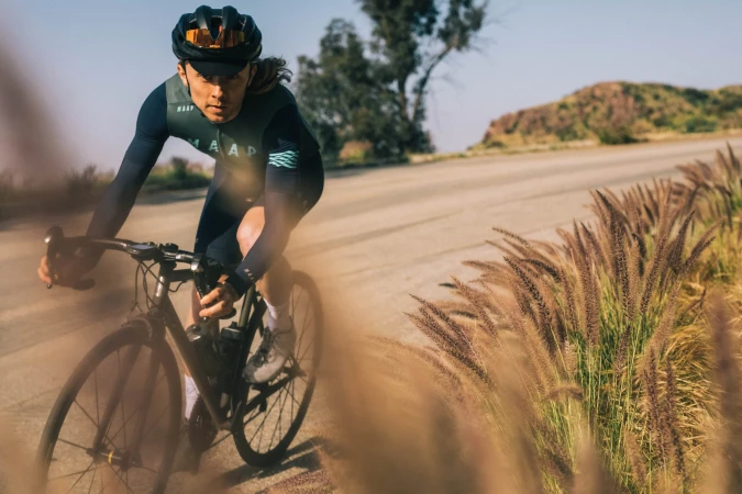 Cyclist in dark green jersey and black helmet riding road bicycle along a paved road past tall grassy weeds.