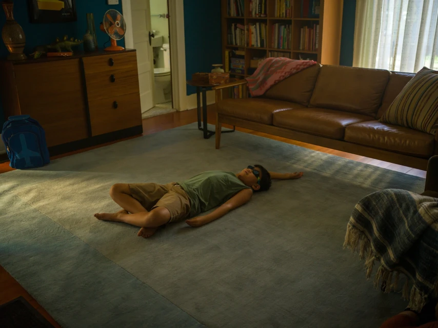 Young child in a green shirt lies barefoot on a blue rug in a living room with a couch, bookcase, and blue wall with cabinet.