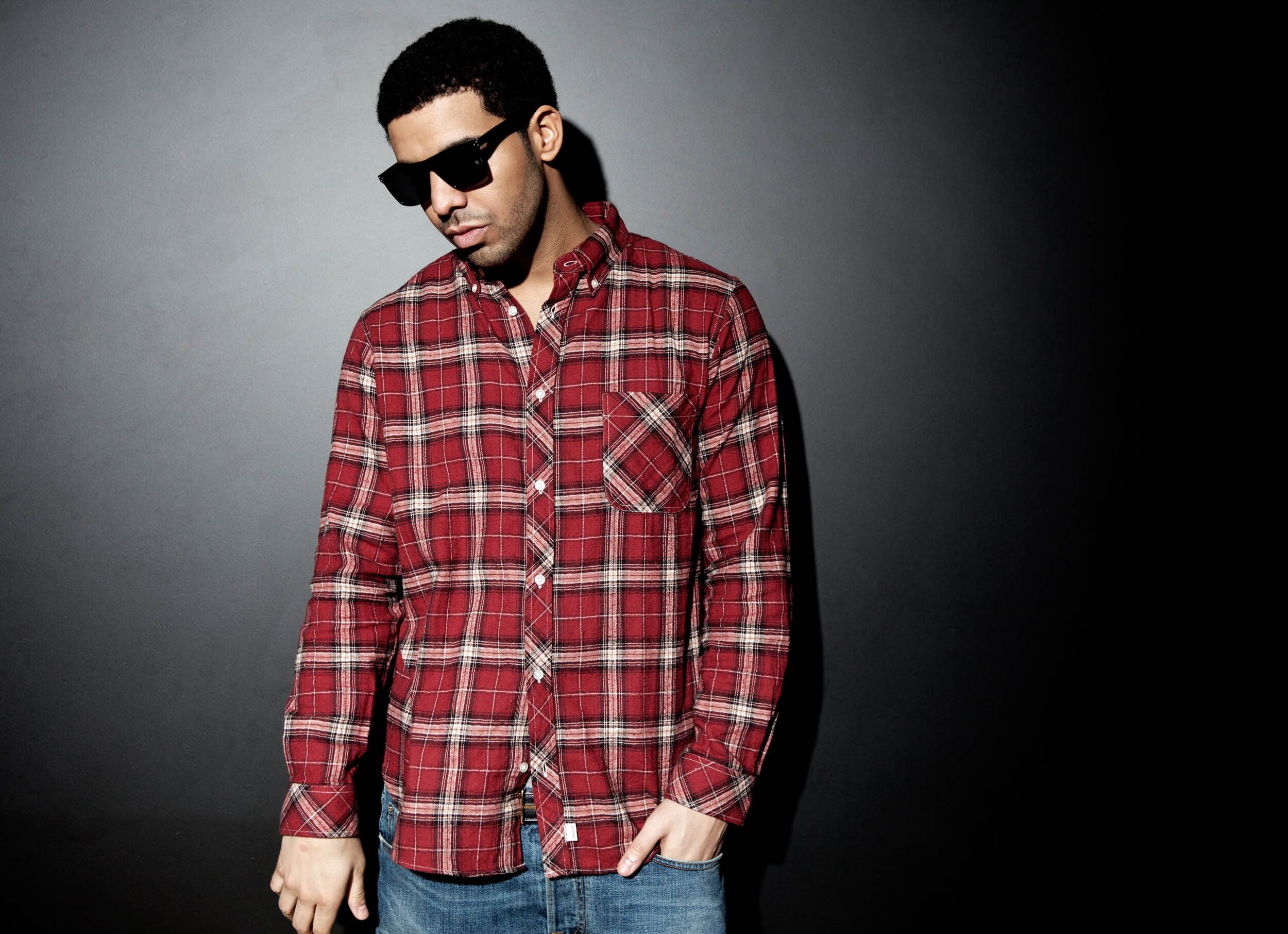 Portrait of Drake wearing sunglasses, a red plaid button-down shirt, and blue jeans against a dark gray gradient background.