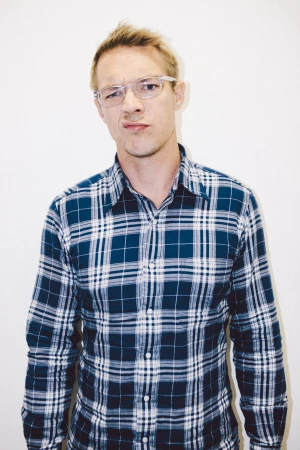 Man wearing blue and white plaid shirt and clear-frame glasses against a plain white background