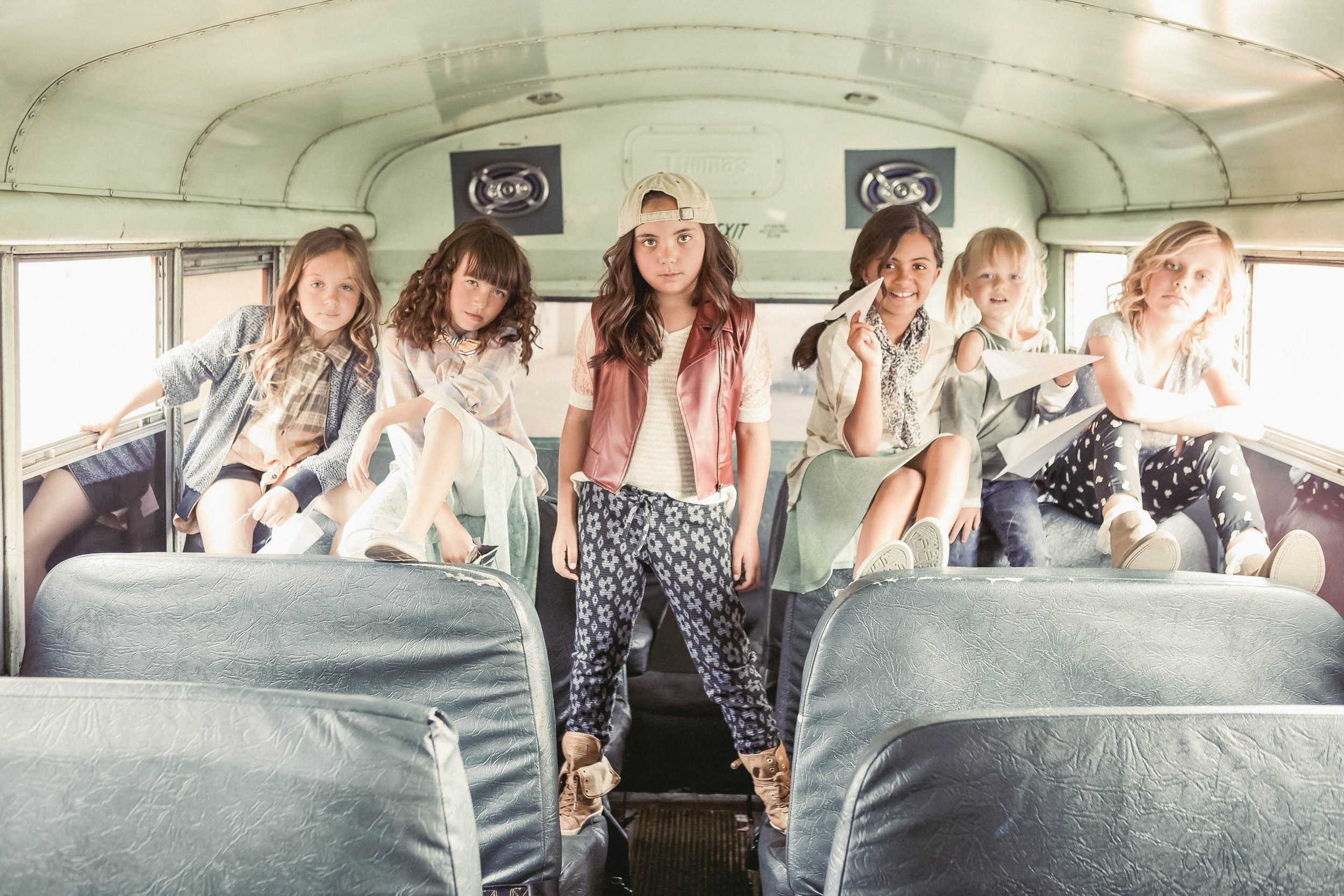 Five young girls inside a vintage bus, dressed in casual fashion, some sitting on seats, one standing, creating a nostalgic and playful scene.