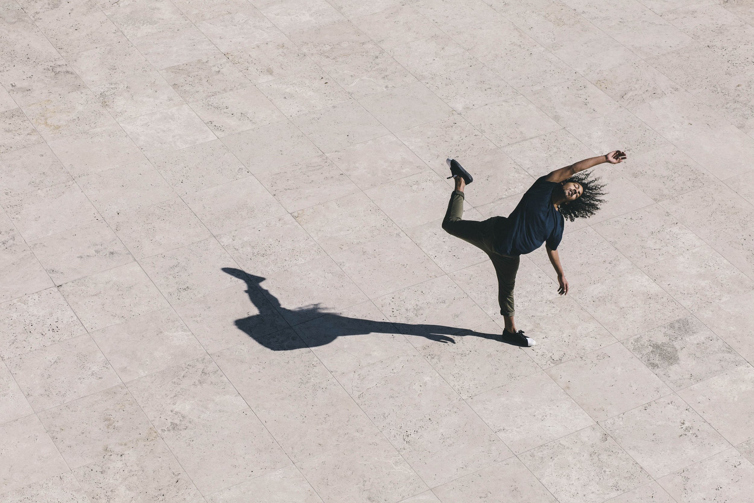 Overhead shot of a person dancing on a light stone tiled surface with long shadows, showing movement and balance in an outdoor setting.