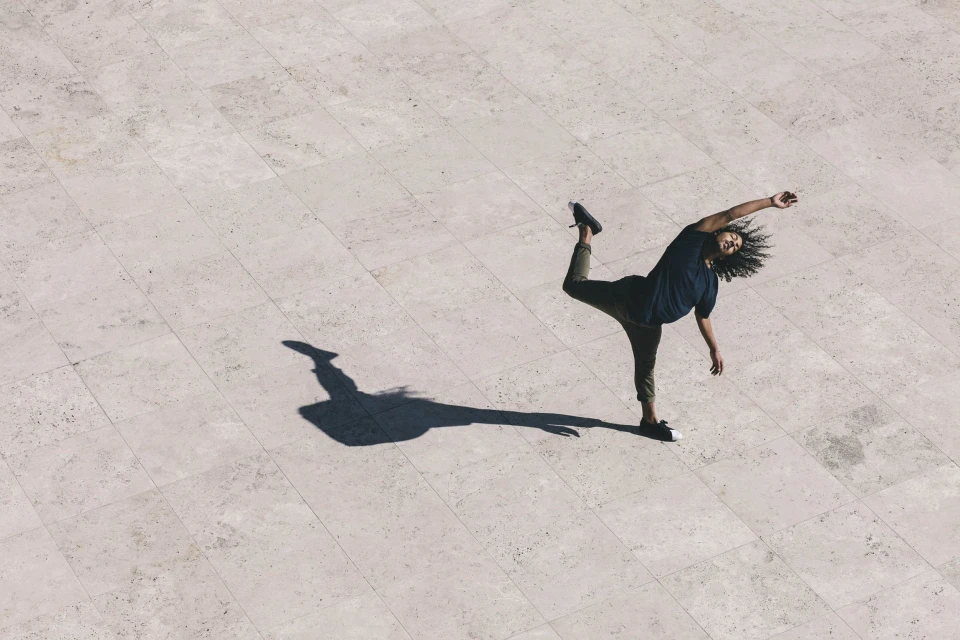 Overhead shot of a person dancing on a light stone tiled surface with long shadows, showing movement and balance in an outdoor setting.