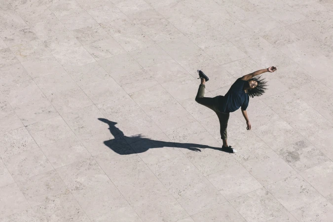 Overhead shot of a person dancing on a light stone tiled surface with long shadows, showing movement and balance in an outdoor setting.