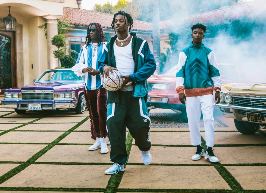 Three stylish men pose with classic cars and a basketball on a patterned stone driveway in front of a Mediterranean-style house.