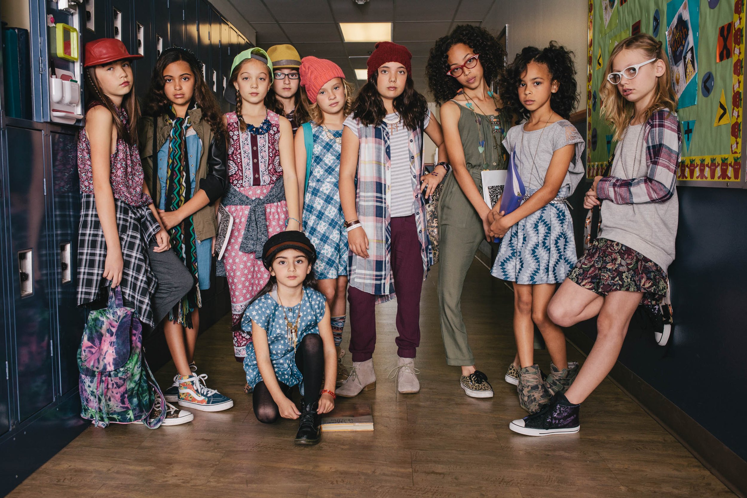 Group of nine young girls in fashionable clothing stand in a school hallway in front of blue lockers. The girls wear hats, glasses and sneakers.