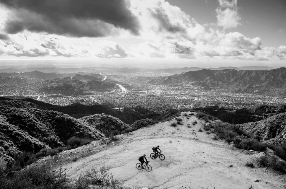 Two cyclists on mountain bikes descend a dirt trail in a wide shot, with a sprawling city and distant mountains in the background on a cloudy day.