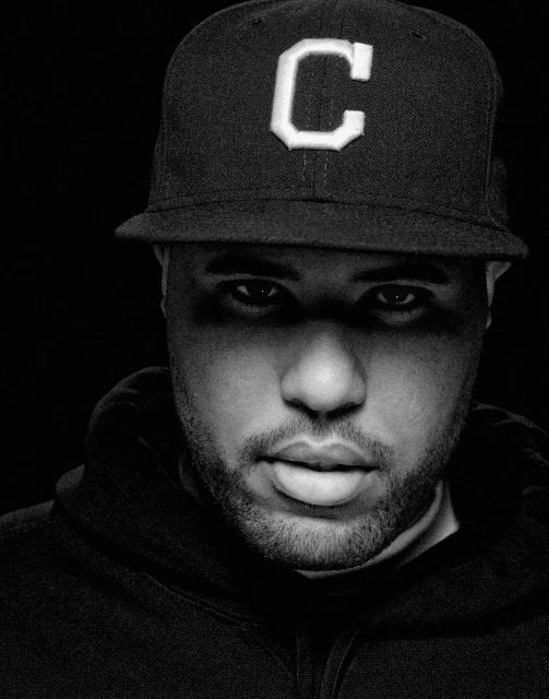 Black and white portrait of a Black adult male wearing a ball cap with a large letter 'C' and a hooded sweatshirt against a dark background.