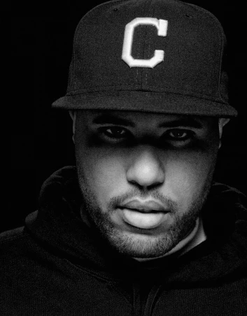 Black and white portrait of a Black adult male wearing a ball cap with a large letter 'C' and a hooded sweatshirt against a dark background.