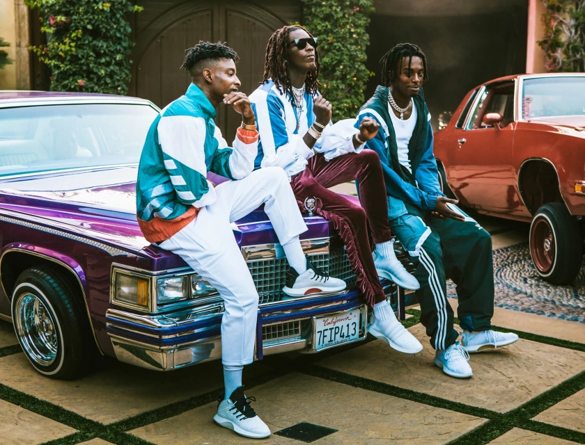 Three adult males pose seated on the hood of a purple vintage car, with a red car visible in the background, in an outdoor setting.