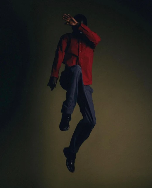 A person is jumping wearing a red, button-down shirt and grey pants against a green background in a dynamic and stylish portrait.