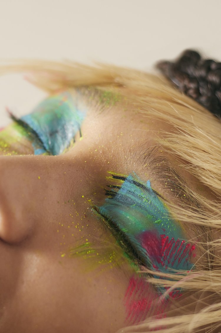 Close-up shot of a model wearing vibrant and colorful eye makeup, featuring turquoise, green, and pink hues, complemented by blond hair.