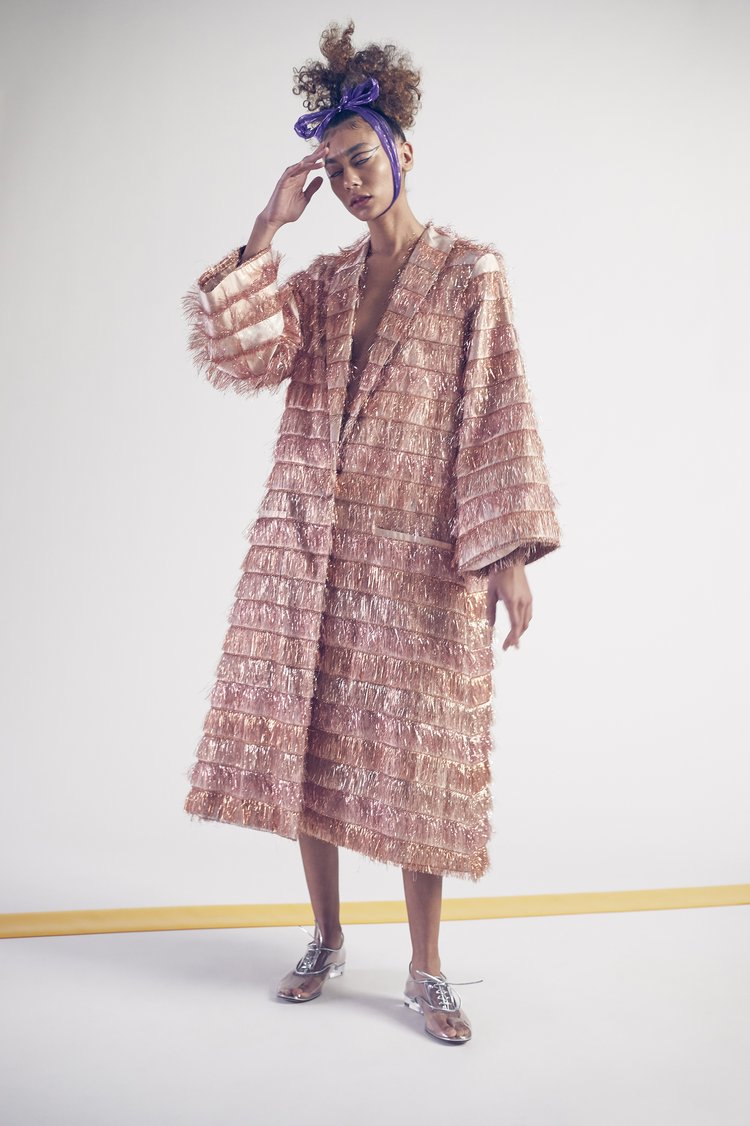 Fashion portrait of a woman in a fringed coat, blue headband, and metallic shoes against a white backdrop with a yellow line.