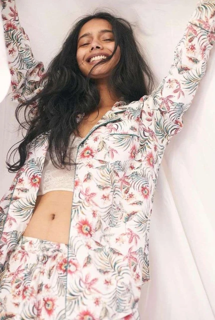 Young woman in floral pajamas smiles with arms raised against a white background, showcasing a relaxed and cheerful indoor scene.