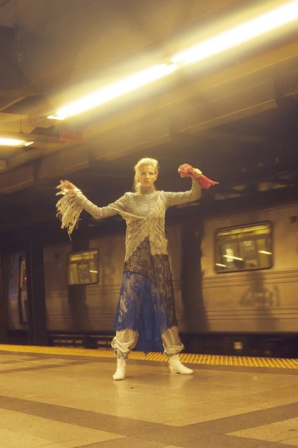 Fashion photo of a woman posing in a unique outfit with white boots, standing on a subway platform with a blurred train in motion.