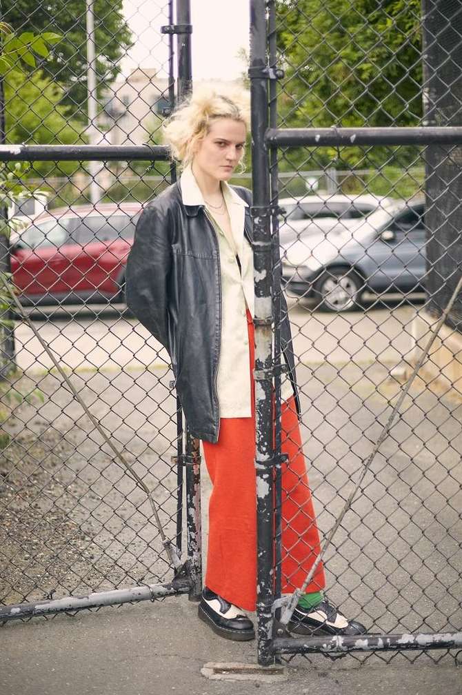 Blonde woman wearing a black leather jacket and red trousers leaning against a chain-link fence outdoors